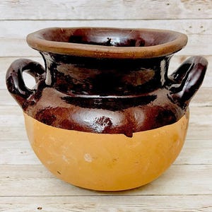 May include: A handcrafted, two-handled earthenware pot with a rounded base and a wide, slightly flared rim. The upper portion is a rich, glossy brown, contrasting with the unglazed, light tan lower half. The pot has a rustic, artisanal aesthetic.