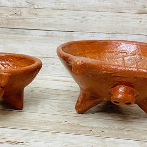 May include: Two terracotta-colored, handmade pig-shaped bowls. The bowls have three legs and a pig snout on the front. The smaller bowl is on the left, and the larger bowl is on the right. The bowls are likely for serving food.