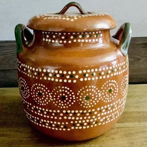 May include: A brown ceramic pot with a lid, featuring white and green dot patterns. The pot has two green handles and a brown handle on the lid. This handcrafted pottery is suitable for kitchen or home decor.