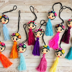 May include: A collection of handmade Frida Kahlo-inspired ornaments. Each ornament features a felt face with embroidered details, a floral headdress, and a colorful tassel. The ornaments are suspended by a black cord, perfect for hanging.