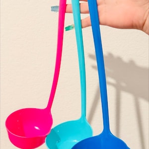 May include: Three plastic soup ladles in bright pink, turquoise, and royal blue. Each ladle features a long, slender handle with a small hole at the top for hanging. The text "Assorted colors shipped out" is visible at the top of the image.