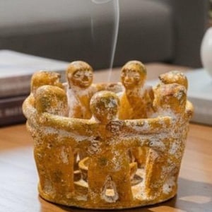 May include: A yellow-brown ceramic incense holder featuring figures in a circle, with smoke rising from the center. The figures are embracing, creating a sense of unity. The holder has a textured, aged appearance.