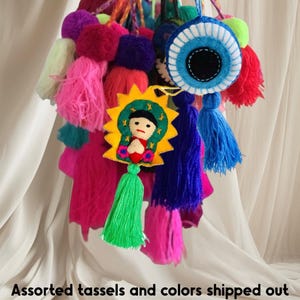 May include: A collection of colorful tassels and pom-poms in various shades of pink, blue, green, purple, and red. Included are felt charms with religious imagery and an evil eye design. The image has text that reads "Assorted tassels and colors shipped out".
