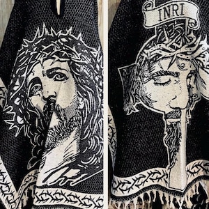 May include: Black and white hooded poncho featuring a detailed illustration of Jesus Christ on the front and back. The back design includes the letters "INRI" on a banner. The poncho has a textured pattern and a fringed hem.
