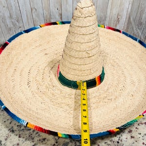 May include: A large, woven straw sombrero with a wide brim and a tall, conical crown. The hat features a colorful trim of blue, red, yellow, and green. The brim measures approximately 10 inches in diameter. The hat is a traditional Mexican accessory.