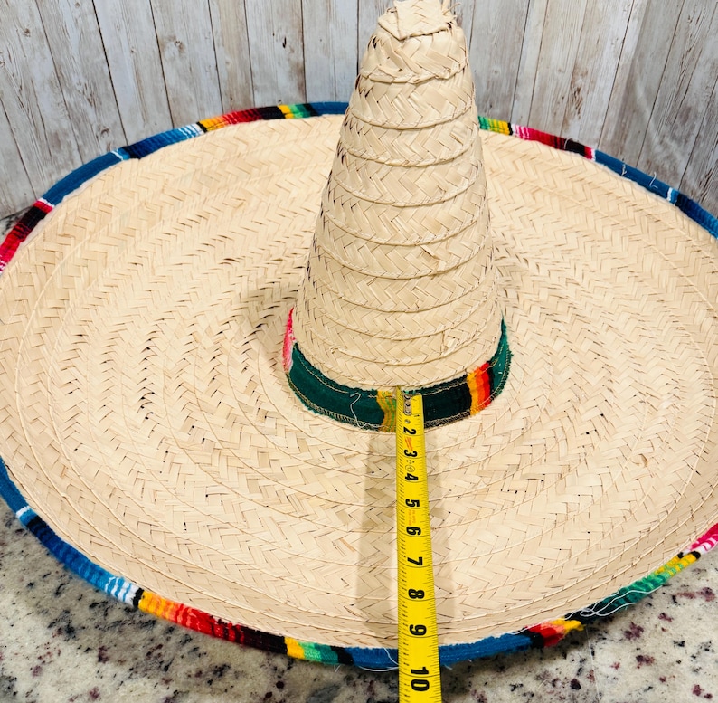 May include: A large, woven straw sombrero with a wide brim and a tall, conical crown. The hat features a colorful trim of blue, red, yellow, and green. The brim measures approximately 10 inches in diameter. The hat is a traditional Mexican accessory.