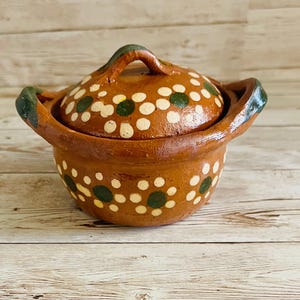 May include: A handcrafted, brown ceramic pot with a lid. The pot and lid are decorated with white and green polka dots. The pot has two handles and a handle on the lid. The pot is sitting on a wooden surface.