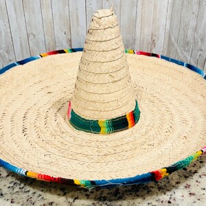 May include: A large, woven straw sombrero with a wide brim and a tall, conical crown. The brim is trimmed with a blue border and colorful woven fabric. The crown features a green band with colorful stripes.
