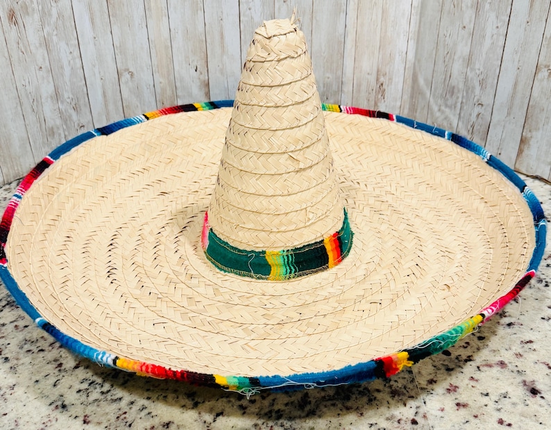 May include: A large, woven straw sombrero with a wide brim and a tall, conical crown. The brim is trimmed with a blue border and colorful woven fabric. The crown features a green band with colorful stripes.
