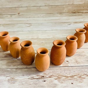 May include: A collection of small, terracotta-colored vases arranged in an arc. Each vase has a rounded body and a flared opening. The vases are displayed on a light-colored, wooden surface.