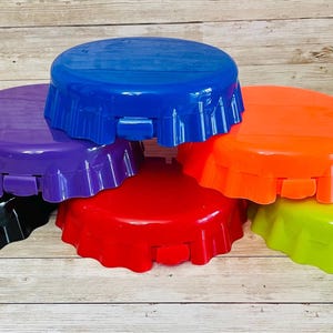 May include: A collection of six colorful bottle cap-shaped items in black, purple, blue, red, orange, and green. The items are arranged in a slightly overlapping pattern, showcasing their vibrant hues and unique design.