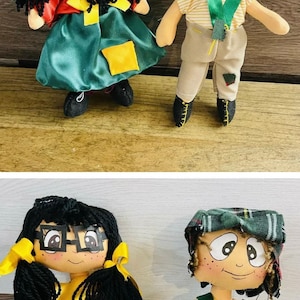 May include: Two handmade dolls, one with black yarn hair, glasses, and a green dress, the other with a plaid hat, freckles, and tan pants. Both dolls have painted faces and are standing on a wooden surface.