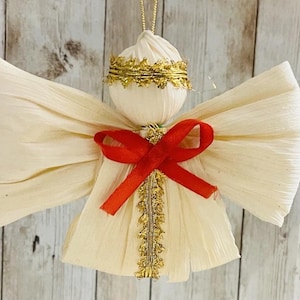 May include: A handcrafted angel ornament with cream-colored wings and body, a red ribbon bow, and gold trim. The angel has a gold halo and is suspended by a gold string. This decorative item is perfect for holiday decor.
