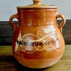 May include: A terracotta-colored ceramic pot with a lid and two handles. The pot features a decorative white and brown swirl design. The pot is sitting on a wooden surface.