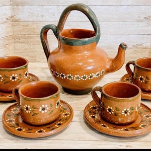 May include: A terracotta tea set featuring a teapot with a handle and spout, and four matching cups and saucers. Each piece is adorned with white floral designs and green accents, creating a rustic aesthetic.