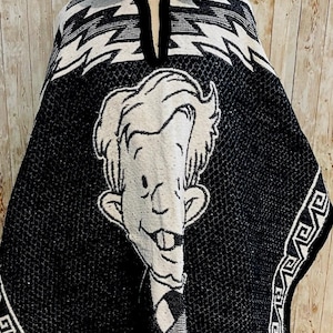 May include: Black and white hooded poncho featuring a cartoon character on the front. The poncho has a geometric pattern on the top and bottom edges. The character has a large head, buck teeth, and a striped tie. The poncho is made of a woven material.