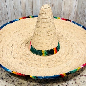 May include: A large, woven straw sombrero with a wide brim and a tall, conical crown. The brim is trimmed with a blue border and colorful woven fabric. The hat features a green band with colorful stripes around the base of the crown.