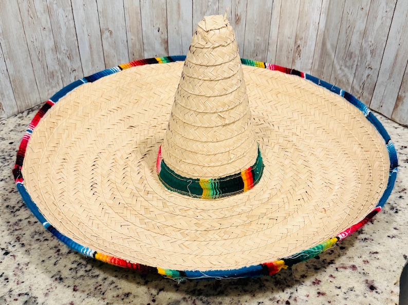 May include: A large, woven straw sombrero with a wide brim and a tall, conical crown. The brim is trimmed with a blue border and colorful woven fabric. The hat features a green band with colorful stripes around the base of the crown.