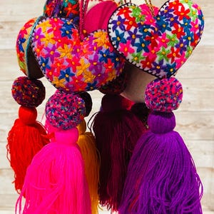 May include: A collection of colorful heart-shaped ornaments with embroidered floral patterns. Each ornament has a tassel in a vibrant color, including red, pink, yellow, and purple. The ornaments are suspended by matching cords.
