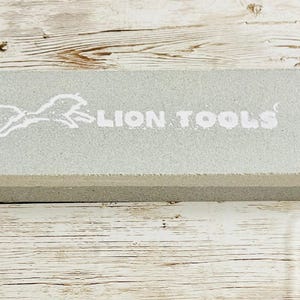 May include: A rectangular, light gray sharpening stone with the words "LION TOOLS" in white. A white lion graphic is to the left of the text. The stone is on a light brown wooden surface.