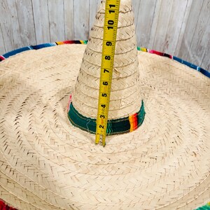 May include: A woven straw sombrero with a colorful trim. The hat's crown measures approximately 11 inches in height. The brim is wide and circular, with a decorative band of green, red, and yellow. The hat is a traditional Mexican style.