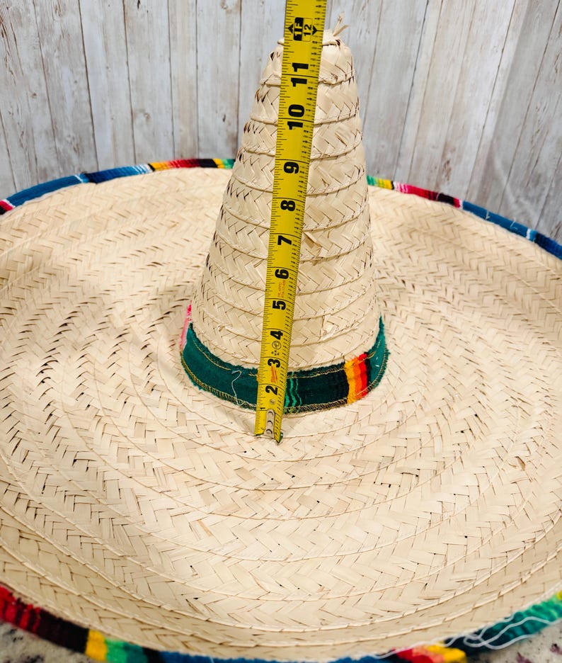 May include: A woven straw sombrero with a colorful trim. The hat's crown measures approximately 11 inches in height. The brim is wide and circular, with a decorative band of green, red, and yellow. The hat is a traditional Mexican style.