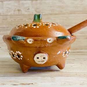 May include: A handcrafted, terracotta-colored ceramic sugar bowl with a lid and a small spoon. The bowl features white and black floral designs, a face-like design, and three small feet. The lid has a green handle.