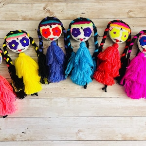 May include: A row of seven Day of the Dead dolls with embroidered faces, braided hair, and colorful yarn tassels. The dolls have different colored eye designs and tassel colors, including pink, yellow, blue, and red.