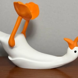 以下が含まれることがあります： オレンジ色のくちばしと上を向いたオレンジ色の足を持つ、白いガチョウの形をしたオブジェ。ガチョウは滑らかなマット仕上げで、小さな黒い目が付いています。長い首が水平に伸び、足が体の上に持ち上げられたデザインです。