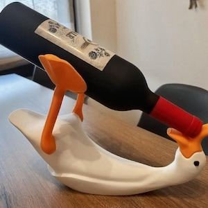 以下が含まれることがあります： 鮮やかなオレンジ色の足とくちばしを持つ白いガチョウ型のワインボトルホルダー。黒いワインボトルを支えており、ボトルには黒い文字が書かれた白いラベルと赤いキャップシールが付いています。ホルダーはボトルを水平に保つように設計されています。