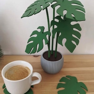 May include: A decorative green monstera plant sculpture with flat, leaf-shaped pieces assembled on thin stems in a ribbed gray pot. A white ceramic mug filled with coffee sits on a green leaf-shaped coaster, with another matching coaster nearby.