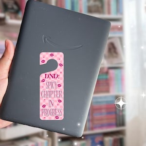 May include: A pink and white door hanger with the text "DND: Spicy Chapter in Progress" and lip graphics is attached to a gray e-reader. The background includes a bookshelf and a window.