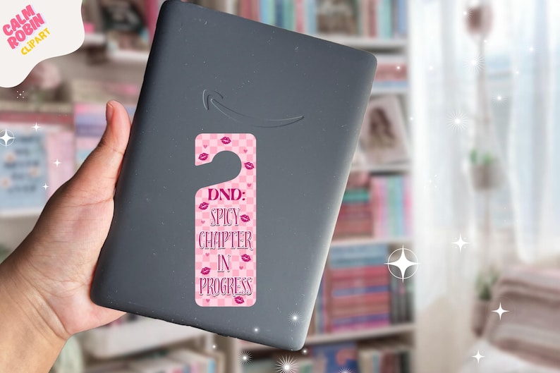 May include: A pink and white door hanger with the text "DND: Spicy Chapter in Progress" and lip graphics is attached to a gray e-reader. The background includes a bookshelf and a window.