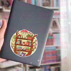 Puede incluir: Un lector de libros electr&oacute;nico negro con una pegatina colorida con un dise&ntilde;o de estanter&iacute;a. La pegatina incluye libros, plantas y setas. La pegatina es redonda y tiene un estilo caprichoso.