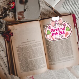 May include: A festive bookmark with the text "Christmas Book Club" is placed in an open book. The bookmark features a pink bow, a snowman, Christmas trees, candy canes, and ornaments. The book has aged pages.
