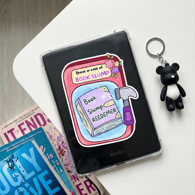 May include: A Kindle e-reader with a clear protective case, featuring a sticker that reads "Break in case of Book Slump." A black and white bear keychain hangs nearby. Books are visible in the background.