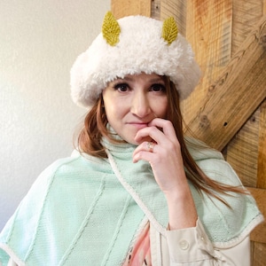 May include: A fluffy white hat with two yellow leaf-shaped accents. The hat is worn with a light green shawl and a beige jacket. The background includes a wooden door and a white wall.