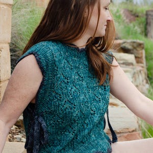 May include: Teal-colored knitted sleeveless top with a detailed openwork pattern. The top has a rounded neckline and a dark blue trim. The garment is made of a soft, textured yarn, perfect for layering or wearing on its own.