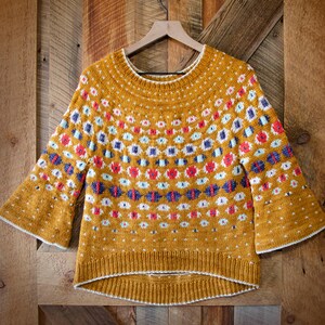 Könnte beinhalten: Senfgelber Strickpullover mit weißem Ausschnitt und Bündchen. Der Pullover hat ein Muster aus roten, blauen und weißen geometrischen Formen. Die Ärmel sind ausgestellt. Der Pullover hängt an einem Holzbügel vor einem Holzgrund.