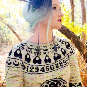 May include: A cream-coloured sweater with black graphic patterns and the numbers 1234567890. The sweater has long sleeves and a round neckline. The person is wearing a black floral headpiece and long, dangling earrings.
