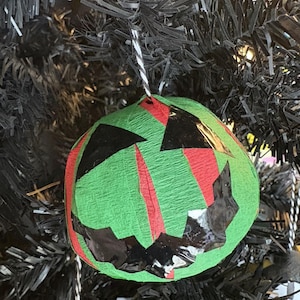 May include: A green, round Halloween bauble with a jack-o'-lantern face. The bauble has black and red accents and is suspended by a black and white string. It hangs on a black artificial tree.