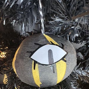 May include: A round, gray and yellow Christmas ornament with a stylized eye design. The ornament is hanging from a black and white string, and is set against a black Christmas tree.
