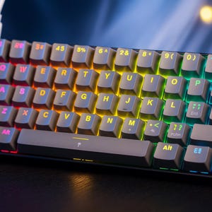 May include: Close-up of a mechanical keyboard with gray keycaps and multi-colored LED backlighting. The keys are arranged in a standard QWERTY layout. The keyboard has a compact design, with a dark gray frame.