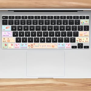 Peut inclure: Clavier d'ordinateur portable avec des autocollants décoratifs. Les autocollants présentent des couleurs pastel et de jolies illustrations de fleurs, d'ours et d'autres motifs. Le texte "have a good day" est visible sur l'un des autocollants. Le clavier est sur une surface en bois.