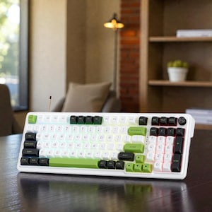Może przedstawiać: Biała klawiatura mechaniczna z zielonymi i czarnymi klawiszami. Klawiatura posiada klawiaturę numeryczną i różne klawisze funkcyjne. Klawisze są podświetlane kolorową poświatą. Klawiatura znajduje się na ciemnej drewnianej powierzchni.