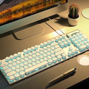 May include: A white, retro-style computer keyboard with round keys and teal backlighting. The keyboard is on a desk with a pen, a small cactus in a white pot, and a computer monitor in the background. The keyboard has a vintage typewriter aesthetic.