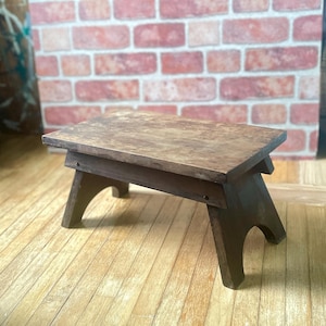 Può includere: Un piccolo sgabello in legno fatto a mano con un piano rettangolare e quattro gambe angolate. Lo sgabello è realizzato in legno tinto scuro, che mostra la venatura naturale. Il design è semplice e rustico, adatto a vari usi.