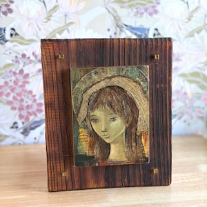 May include: A framed artwork featuring a portrait of a person with long hair and a halo. The portrait is set within a rectangular frame made of dark brown wood, with brass-colored accents. The artwork has a vintage aesthetic.