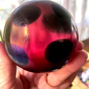 May include: A round, glass orb with a vibrant red and purple hue, adorned with large, irregular black spots. The orb's surface reflects light, creating a shimmering effect. The object is held in a person's hand.