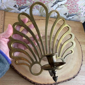 May include: A brass wall sconce with a fan-shaped design. The sconce features a candle holder and a decorative, openwork pattern. The brass has a warm, golden hue. The sconce is designed to be mounted on a wall and is suitable for holding a single candle.